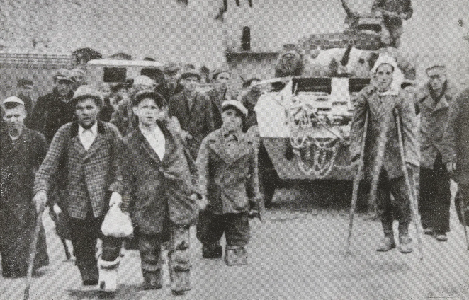 Photo of prisoners. Some are walking with crutches. Behind them is a tank.